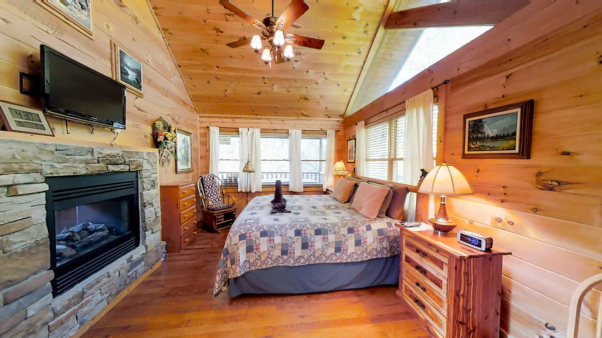 A serene master bedroom featuring a plush king bed positioned near a two-sided seasonal fireplace. Natural light pours in through ample windows with woodsy views, while warm wooden furniture and a ceiling fan add comfort. The tranquil scene is completed by soft linens and thoughtful decorative touches, evoking a restful and romantic atmosphere.