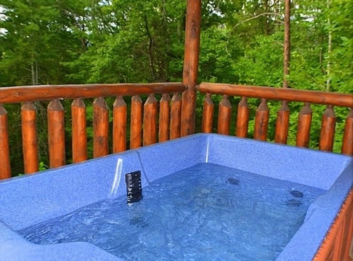 A cozy outdoor hot tub sits on a wooden patio, surrounded by lush greenery. The wooden railing frames an inviting space perfect for soaking while immersed in nature. The atmosphere is serene, ideal for relaxation and conversation under the stars.