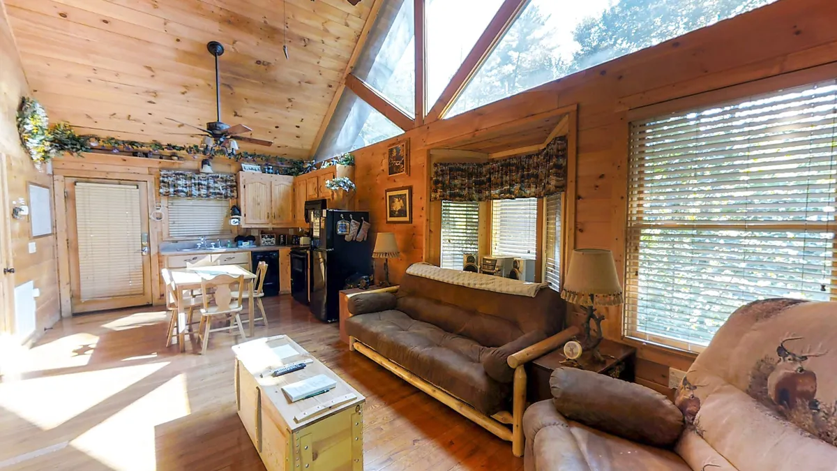 A wide shot of an open-plan living space filled with natural light from large windows, showcasing rustic wooden furniture and homey decor. The kitchen area is visible in the background, while the dining table and chairs are placed in a sunlit spot, perfect for enjoying meals together.