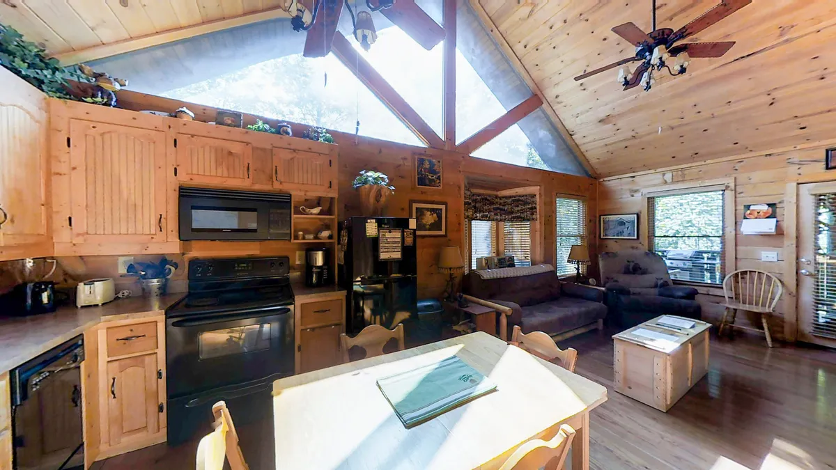 This image captures an open layout that blends the kitchen and dining spaces with a central table, wooden cabinetry, and comfortable seating. Large windows in the background bring in ample natural light, completing the cozy and inclusive environment ideal for socializing and sharing meals.