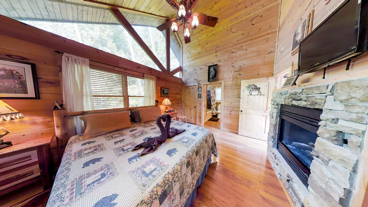 This image features a cozy interior with a bed adorned with a patterned quilt, a rustic stone fireplace, and expansive windows letting in abundant natural light. The warm glow from a chandelier enhances the intimate ambiance, creating a perfect setting for quiet moments spent together. The scene conveys comfort and togetherness, with opportunities for snuggling by the fire or sharing a morning coffee while watching the sun rise over the tranquil landscape outside.