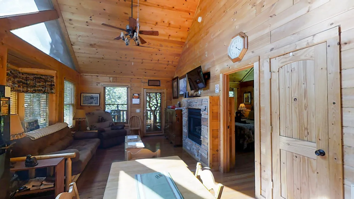 A bright, inviting living area with exposed wooden beams, a stone fireplace, ample seating, large windows letting in natural light, and a dining table. The view outside emphasizes the cozy retreat aspect while promising beautiful scenery year-round.