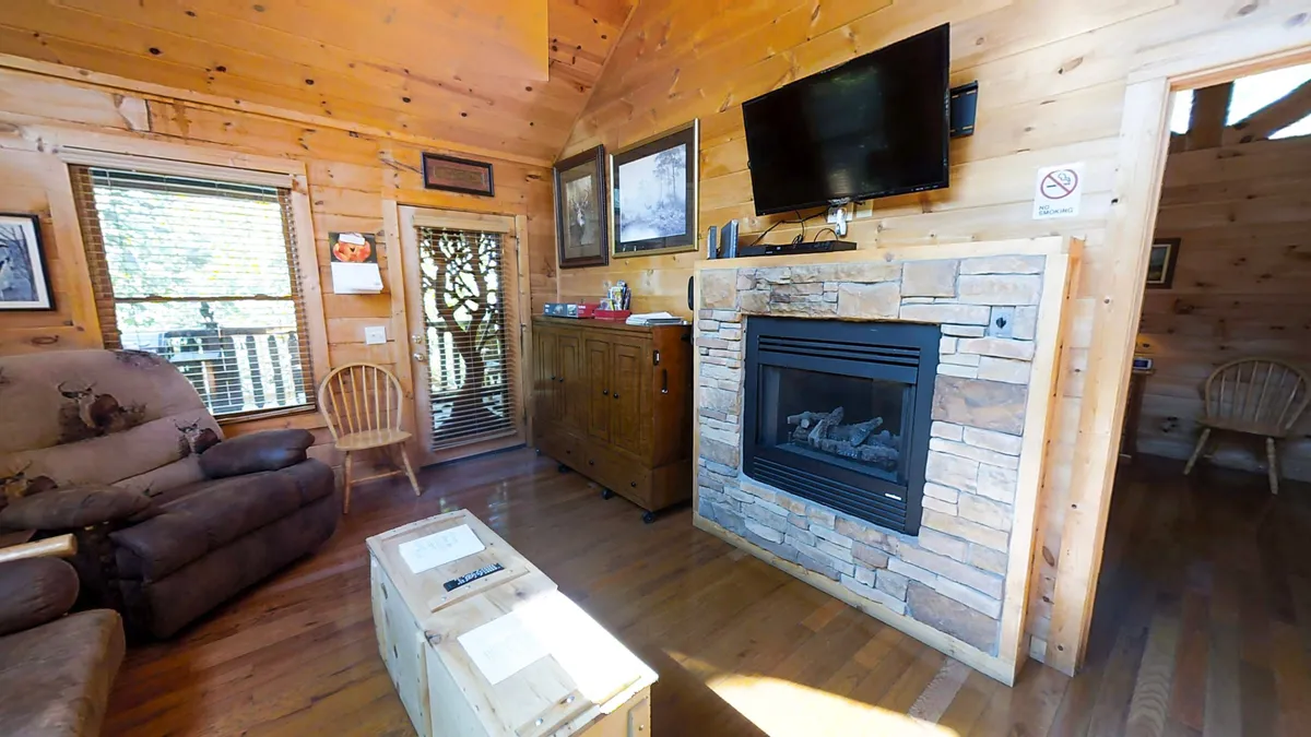 This image presents a cozy living room anchored by a substantial stone fireplace, surrounded by comfortable seating and rustic wooden furniture. A television is clearly visible, and the arrangement encourages relaxing, watching movies, and socializing. Warm sunlight streams through the windows, enhancing the inviting atmosphere.