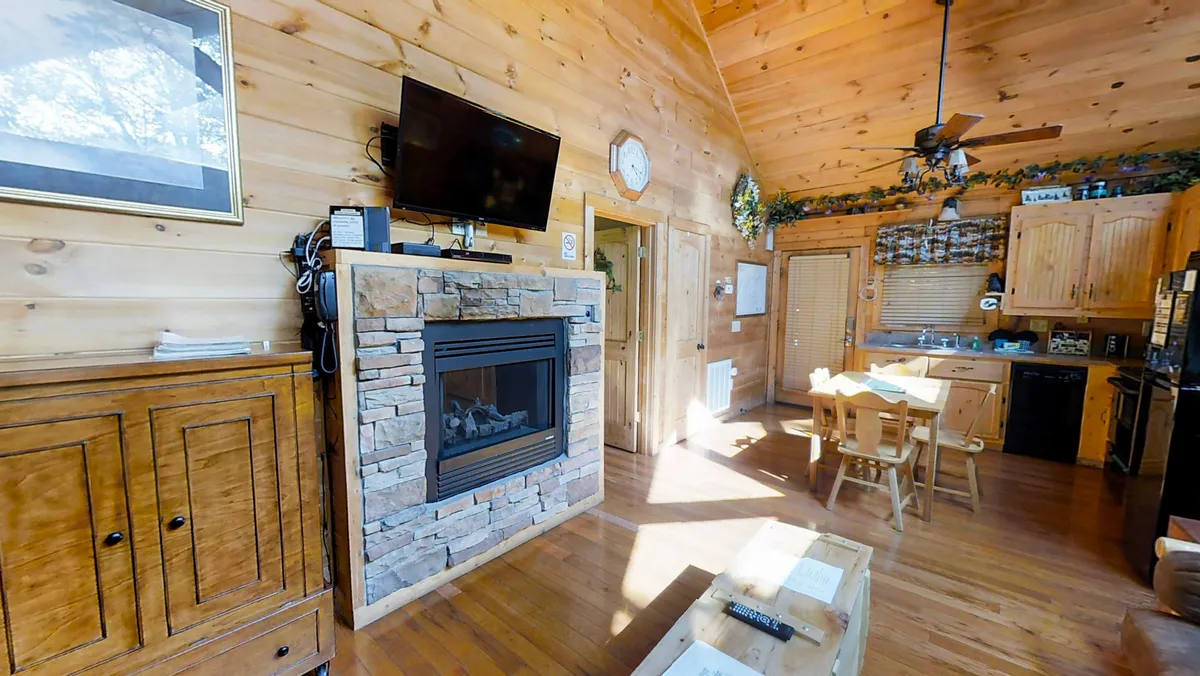The scene captures an open interior layout that blends a rustic stone fireplace, a dining area, and a kitchen space with wooden cabinetry. There's visible seating around the fireplace, and a TV is in view, suggesting the space is used for both family gatherings and relaxing evenings.